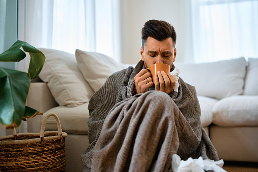 Man with flu virus drinking hot tea while sitting wrapped in a blanket at home.