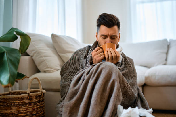Sick man wrapped in blanket drinking tea at home. Man with flu virus drinking hot tea while sitting wrapped in a blanket at home. immune system stock pictures, royalty-free photos & images