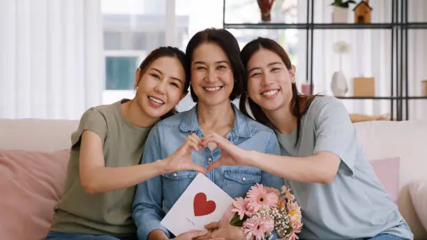 Photo of Enjoy good warm time Mother day two grown up kid child girl look at camera joy cuddle hug make heart shape love hand sign to mature mum. Asia middle age mom young adult three people smile happy face