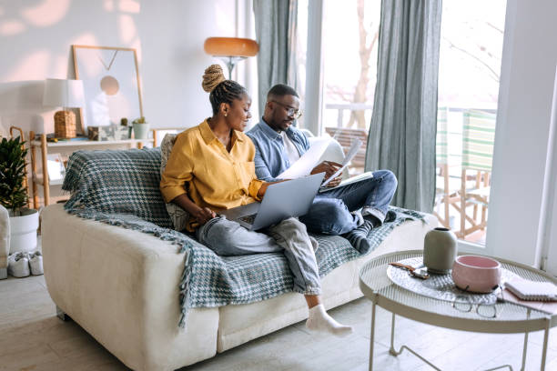 jeune couple souriant travaillant à domicile, passant en revue la paperasse - laptop sofa men computer photos et images de collection