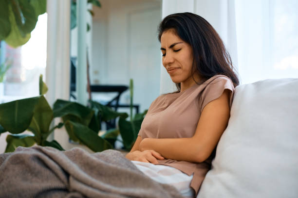 Woman with stomach pain on the sofa at home. Mid adult woman suffering from stomach pain while sitting on sofa in the living room. endometriosis stock pictures, royalty-free photos & images