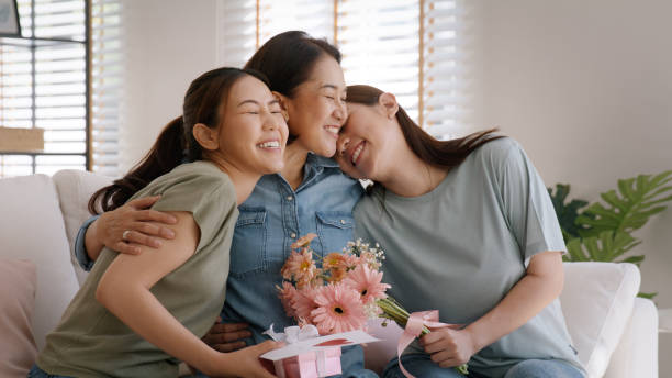 festa della mamma due bambino adulto coccola abbraccio dare fiore confezione regalo rosso cuore carta alla mamma matura. amore bacio cura mamma asia mezza età adulto tre persone sedute a casa divano felice sorriso godere di tempo in famiglia. - mothers day mother kissing child foto e immagini stock