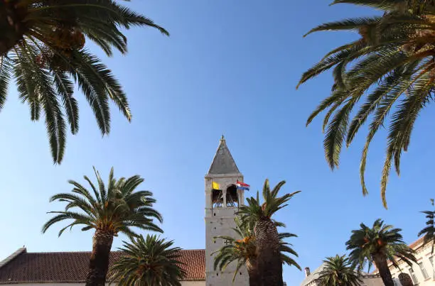 Old church with a tiled roof and a high stone bell tower among green palm trees on a sunny summer day