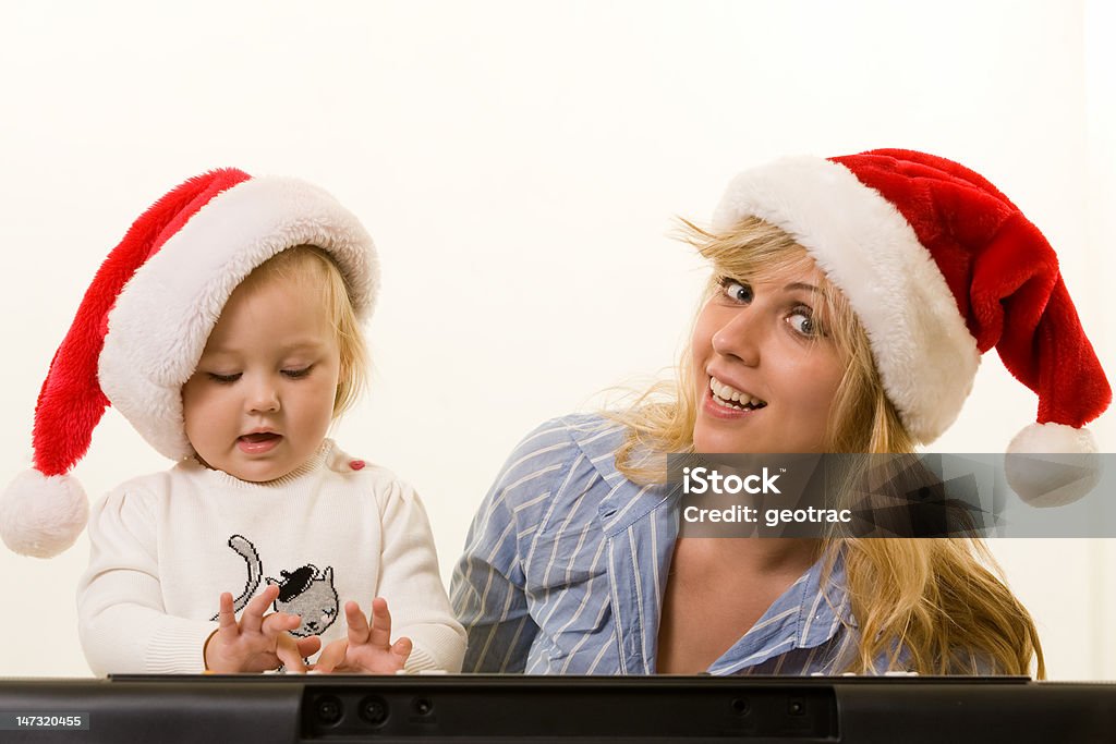 Christmas carols Adorable caucasian blond baby girl toddler sitting with mom both wearing Santa caps in front of a keyboard Adult Stock Photo