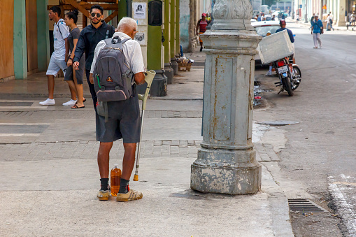 Havana, Cuba - January 20, 2023: Rear view of a senior man with a knapsack and a walking stick standing on the side of a city street. Vehicles and buildings are in the background; incidental people are walking along the road.