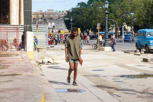 Havana, Cuba - January 23, 2023: Man walking down a sidewalk on a sunny day in shorts and sneakers. Vehicles, trees, buildings, and incidental people are in the background.