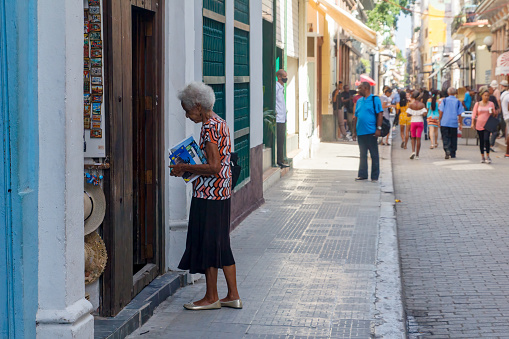 Havana, Cuba - January 25, 2023: A senior woman holding some books and standing by the entrance to a street-side store. Buildings line both sides of the road; incidental people are walking along the street.