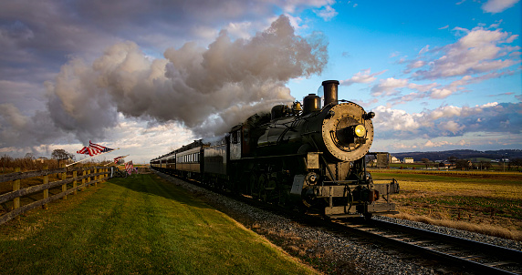 A View of a Classic Steam Passenger Train Approaching, With American Flags Attached to a Fence on a Sunny Autumn Day