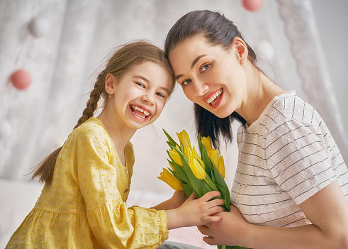 Happy women's day! Child daughter is congratulating mom and giving her flowers tulips. Mum and girl smiling and hugging. Family holiday and togetherness.