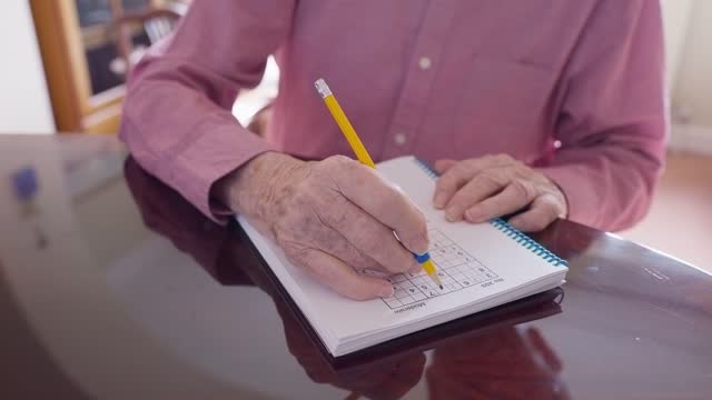 Close Up Of Senior Man With Arthritis Using Grip Aid On Pencil To Write In Sudoku Puzzle Book