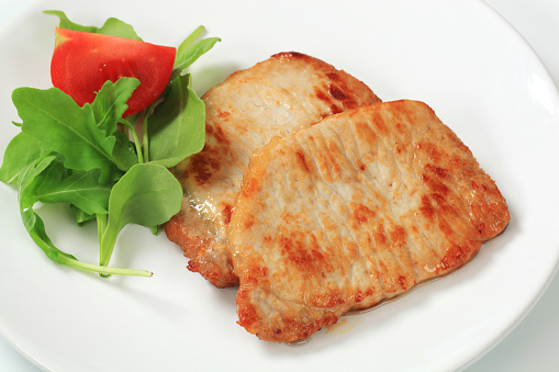 Two roasted pork steaks, green salad with arugola and tomato on a white plate