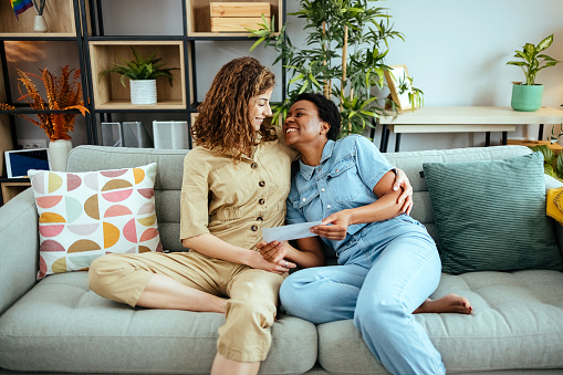 Smiling multiracial lesbian pregnant couple looking baby ultrasounds at home sitting on the couch