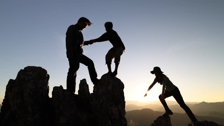 silhouette of helping hands during climbers A man helps a man climb a steep rock and has people helping and teamwork concept. Shadow on the mountain with sunlight. Success on the cliff.