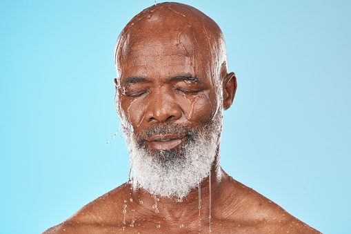 Black man, face and water drip on studio blue background for wellness, healthy skincare and bathroom hygiene. Male model, wet shower and water drops for mature beauty, facial and fresh body cleaning