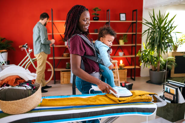 Mother at home ironing clothes with baby in sling