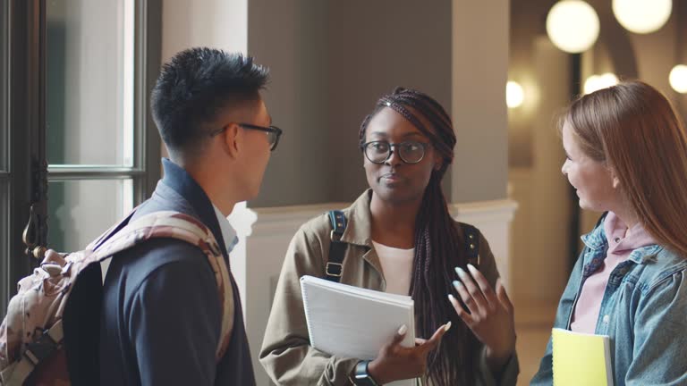 Diverse classmates discussing something while standing in college corridor. Realtime