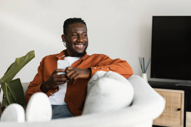 istock Excited black guy chatting on cellphone or surfing internet, sitting on sofa at home, looking aside at copy space 1473237519