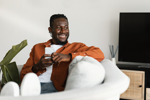 Excited black guy chatting on cellphone or surfing internet, sitting on sofa at home, looking aside at copy space and smiling. Modern technologies and lifestyle