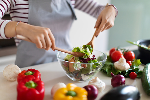 Cooking concept. Unrecognizable woman in grey apron chef making fresh organic vegetable salad, cropped of lady preparing delicious healthy meal at home, mixing veggies in bowl, closeup, copy space