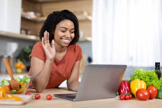 istock Smiling young black lady in red t-shirt preparing lunch, waving hand at computer webcam, shooting food blog 1473237704