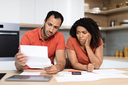 Upset young black woman and man in red t-shirts work with documents check finance bills, suffers from stress, crisis in kitchen interior. Problems with debts, credit and mortgage, bookkeeping at home