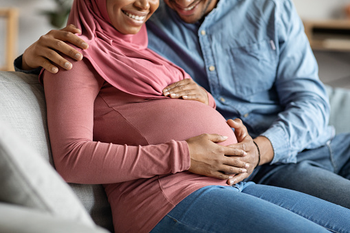 Black pregnant muslim couple relaxing together on couch at home, happy islamic family awaiting for a baby tenderly touching belly and smiling, lady in hijab and her husband enjoying future parenthood