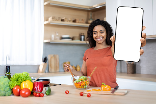 Cheerful millennial black lady cuts organic vegetables at salad, shows smartphone with blank screen in kitchen interior. App for food blog, social networks, household chores, cooking dinner at home