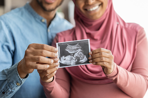 Happy pregnant black muslim spouses demonstrating baby sonography photo at camera while relaxing together at home, excited islamic spouses showing ultrasound scan of their child, cropped, closeup