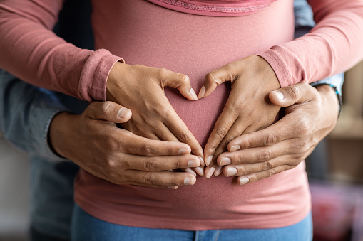 Black muslim couple making heart shape on pregnant belly with their hands, closeup shot of unrecognizable islamic spouses awaiting for a baby bonding at home, making love gesture with arms on tummy