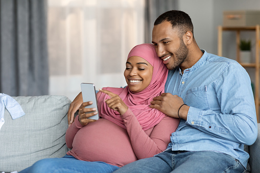 Happy Pregnant Black Islamic Couple Using Smartphone While Relaxing Together At Home, Expectant Muslim Spouses Resting On Couch With Mobile Phone, Browsing Internet Or Shopping Online, Copy Space