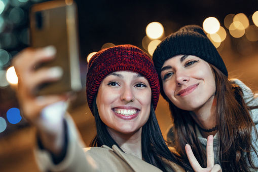 Shot of two joyful woman taking a selfie with smartphone while showing peace gesture in the city at night