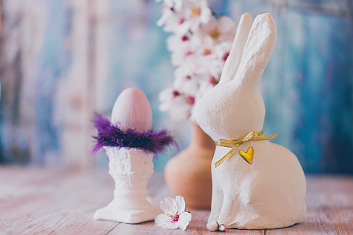 Sweet Easter still lief of one purple painted Easter egg with purple feathers on top of a small white pillar, a branch of almond blossoms and a white Easter bunny on blue vintage background. Creative color editing with added grain. Very soft and selective focus. Part of a series.
