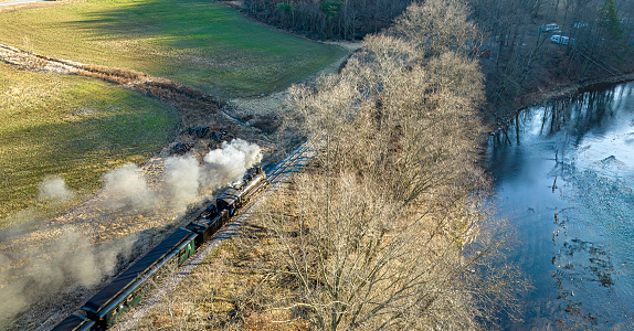 A Behind Aerial View of a Narrow Gauge Steam Passenger Train Traveling Thru Farmlands and a River Blowing Smoke on a Winter Day