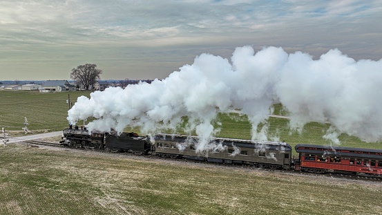 A Side Aerial View of a Steam Passenger Train Approaching, Traveling Thru Open Farmlands, Blowing Lots of White Smoke, on a Winter Day