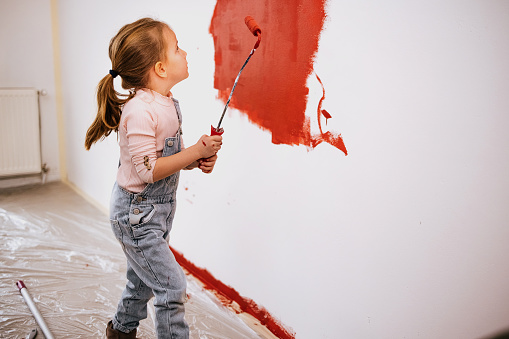 Happy cute child girl painting the wall with paint roller standing in a bright room