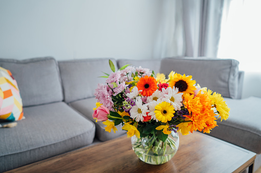 Vase with huge multicolor various flower bouquet on the coffee table with blurred background of modern cozy light living room with gray couch sofa. Giving flowers. Mother's day, birthday gift.