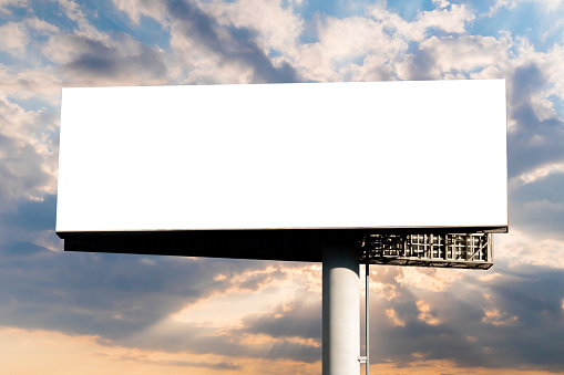 Blank wide white billboard against warm sunset sky. Sun beams shining through dramatic clouds - mock up. White screen, consumerism, advertising, template, mockup and copy space concept