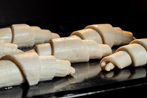 Six uncooked croissants on tray in electric oven - close up view, black background. French cuisine, homemade bakery, breakfast, food, cooking, pastry, semi-finished products and raw concept