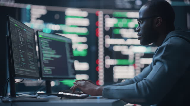 Dolly Shot of a Black Male Programmer Working in a Monitoring Control Room, Surrounded by Big Screens Displaying Lines of Programming Language Code. Portrait of a Man Creating a Software and Coding