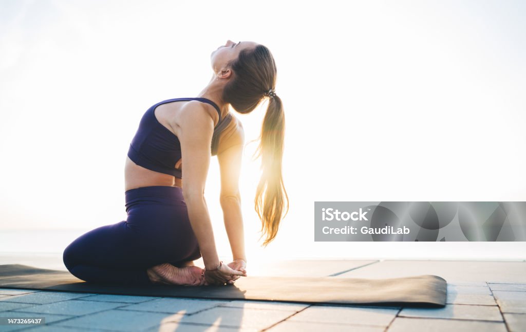 Sportswoman stretching body on quay in sunshine Side view of flexible barefoot lady performing Thunderbolt pose with hands together behind back and bending back during workout on beach Yoga Stock Photo