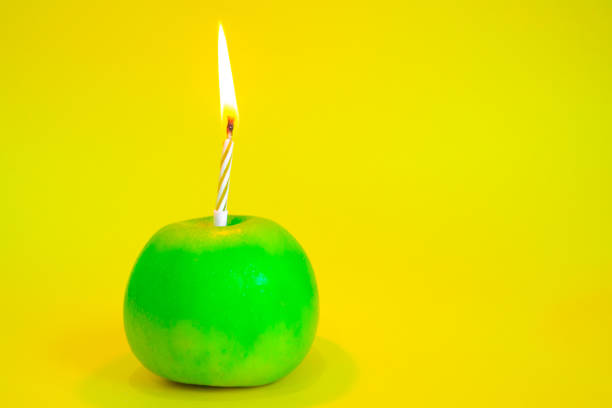 A candle burns in a green apple instead of a cake on a yellow background. Concept of a healthy food, birthday cake with no sugar