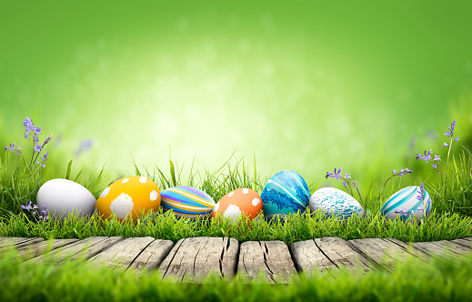 A collection of painted easter eggs celebrating a Happy Easter on a spring day with green grass meadow background with copy space and a rustic woodern bench to display products.