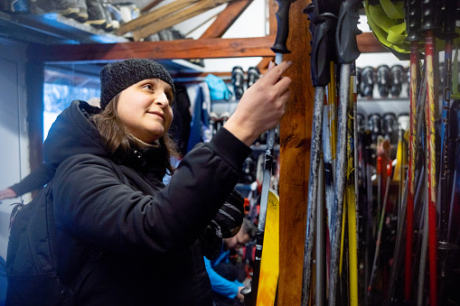 Cheerful female customer in black jacket and black helmet chooses skis at ski rental depot and ski outfitter at ski resort.