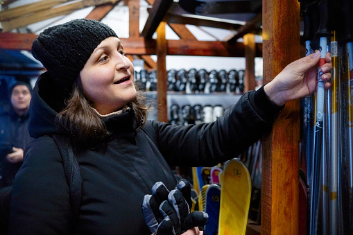 Cheerful female customer in black jacket and black helmet chooses skis at ski rental depot and ski outfitter at ski resort.