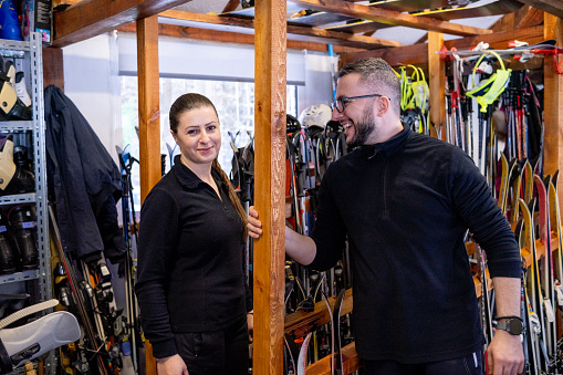 Portrait of two cheerful assistants at a ski and ski equipment rental depot. Skis and poles can be seen behind them.