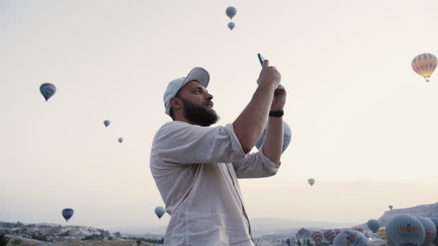 Low angle happy young smiling Caucasian tourist man takes photos of incredible hot air balloons sky view panorama.