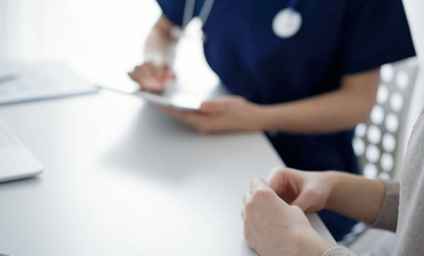 istock Doctor and patient sitting at the table in clinic while discussing something and using tablet computer. The focus is on female patient's hands, close up. Medicine concept 1473276927