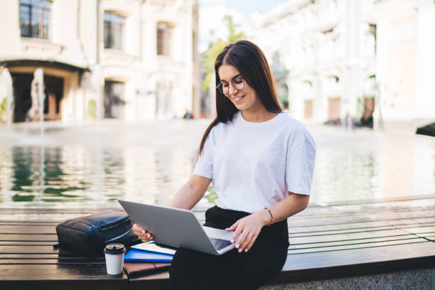 istock Cheerful hipster girl in optical eyewear checking received email message on modern netbook technology, happy digital nomad reading blog publication using public internet connection in city 1473282484