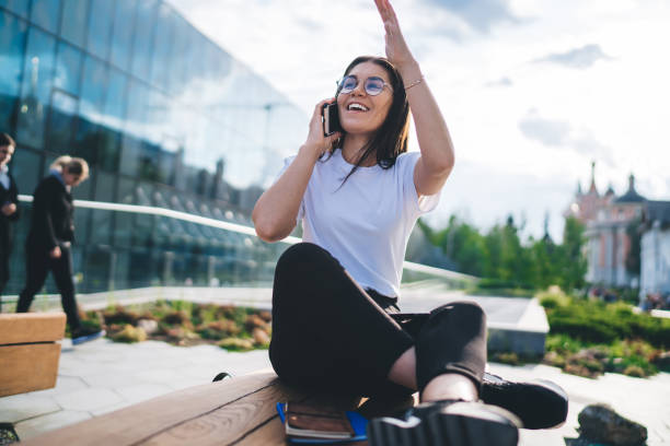 istock Cheerful female student in optical spectacles for eyes correction satisfied with friendly mobile conversation sitting at street urbanity and waving, funny hipster girl using roaming for cell calling 1473282515