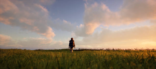istock Little boy with backpack walks in pasture in countryside under a blue cloudy sky during sunset. 3D rendering. 1473282751